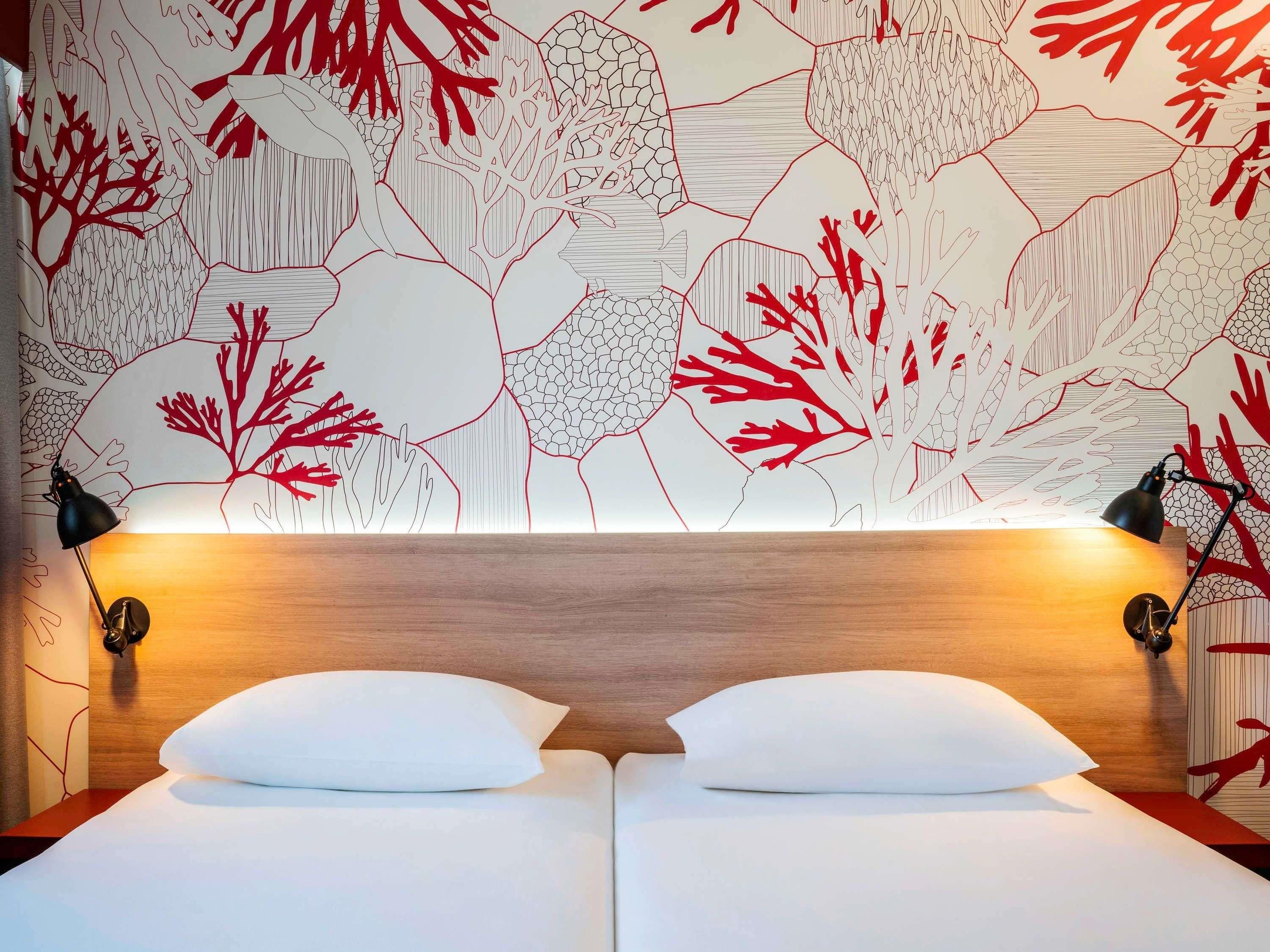 A hotel room with a wooden headboard, two white beds with pillows, and a wall with a red and white abstract pattern.