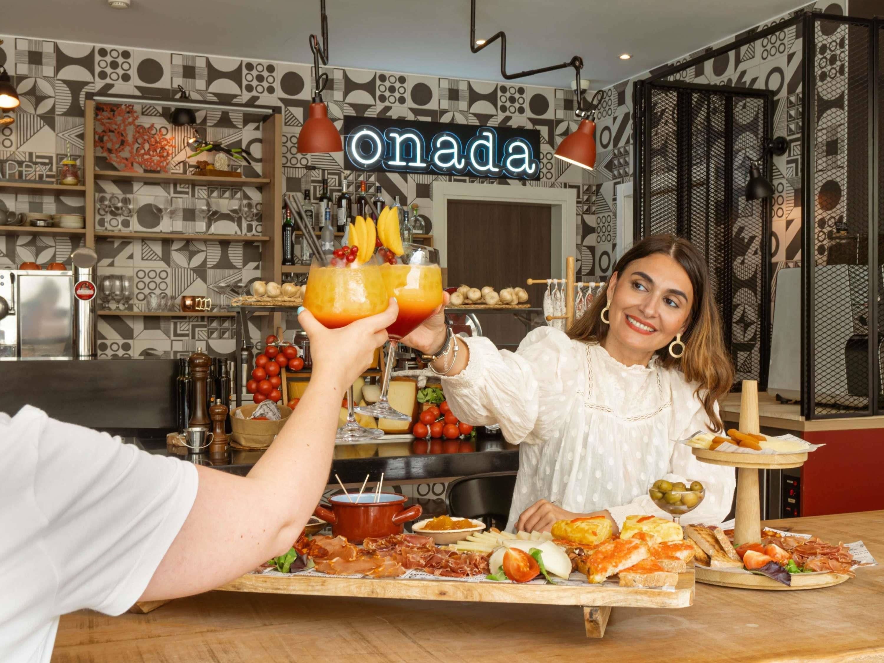 En restauranginteriör med ett modernt, geometriskt mönster på väggen, en neonskylt med texten ”onada” och ett urval av mat- och dryckesalternativ presenterade på en träbricka.