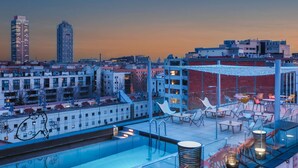 Un área de piscina en la azotea con sillones y mesas, rodeada de edificios y un paisaje urbano al anochecer.