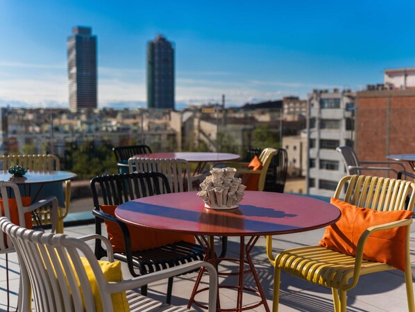 An outdoor seating area with round tables and chairs, a decorative vase, and a cityscape view in the background. - ibis Styles Barcelona City Bogatell (Barcelona)