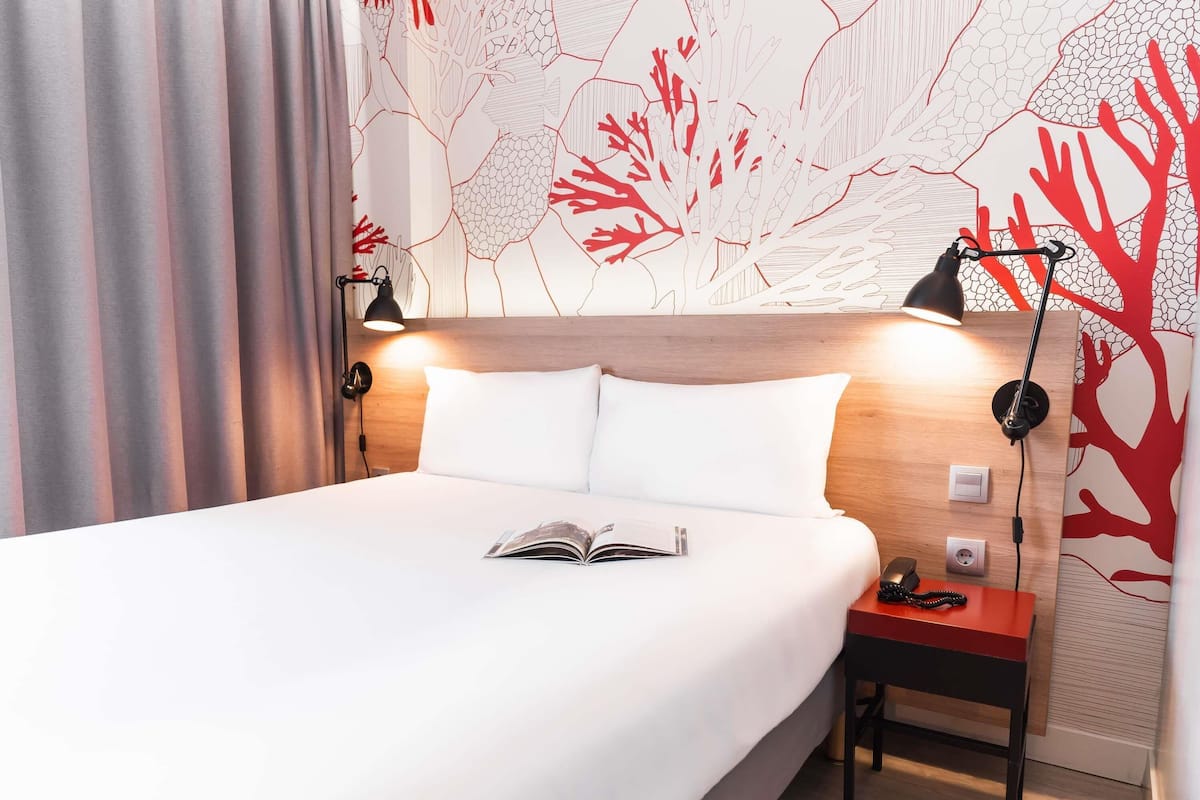 A neatly made bed with a book, a bedside table with a lamp, and a wall with a tree pattern.