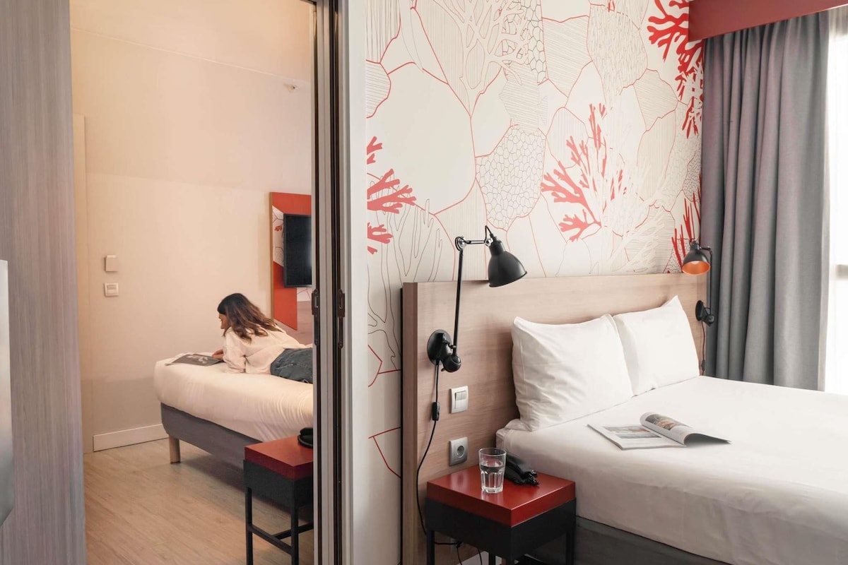 Une chambre d’hôtel comprenant un lit, des tables de chevet, un bureau, une chaise, un téléviseur et un mur orné d’un motif abstrait rouge.