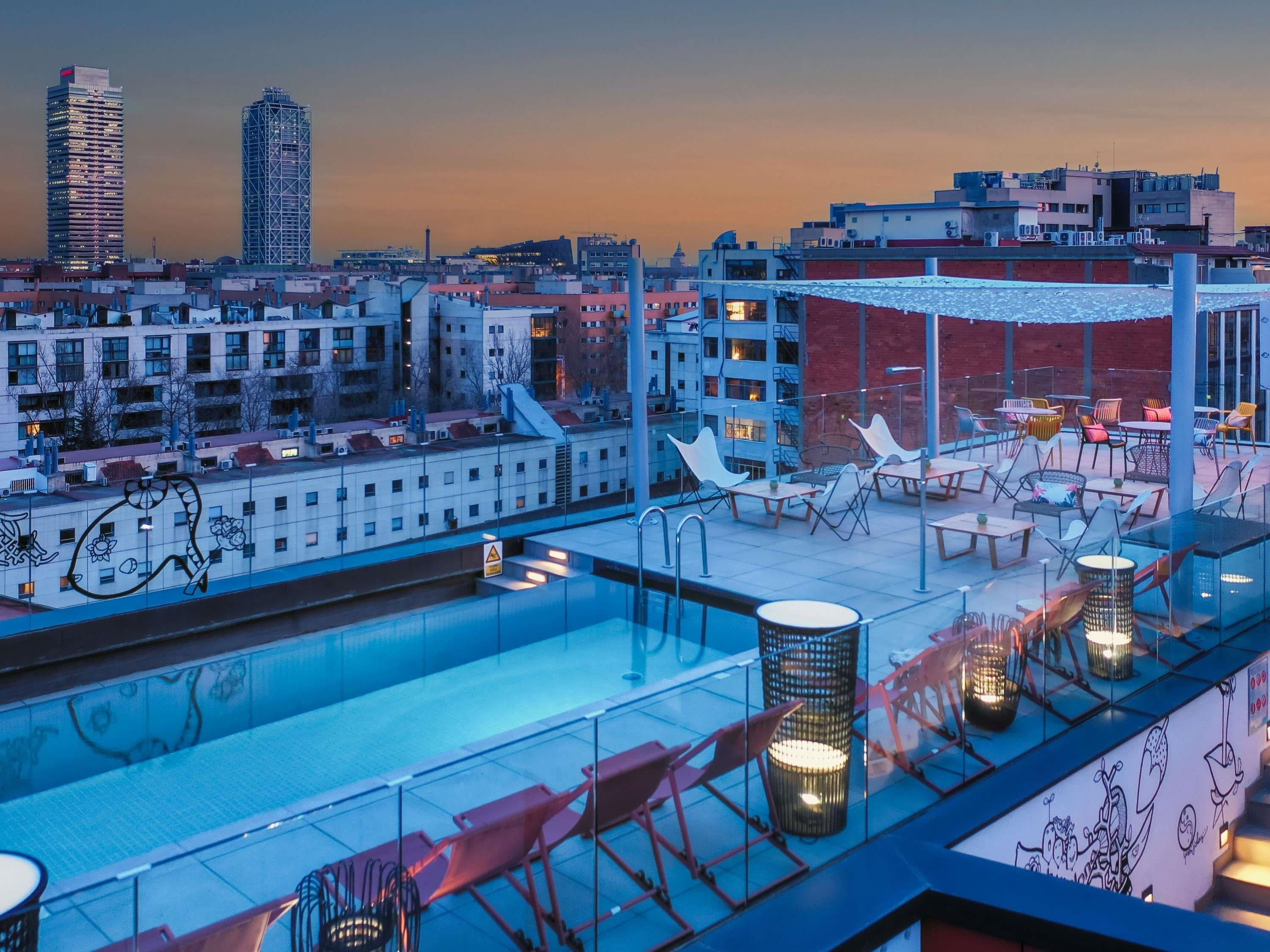 Un área de piscina en la azotea con sillones y mesas, rodeada de edificios.