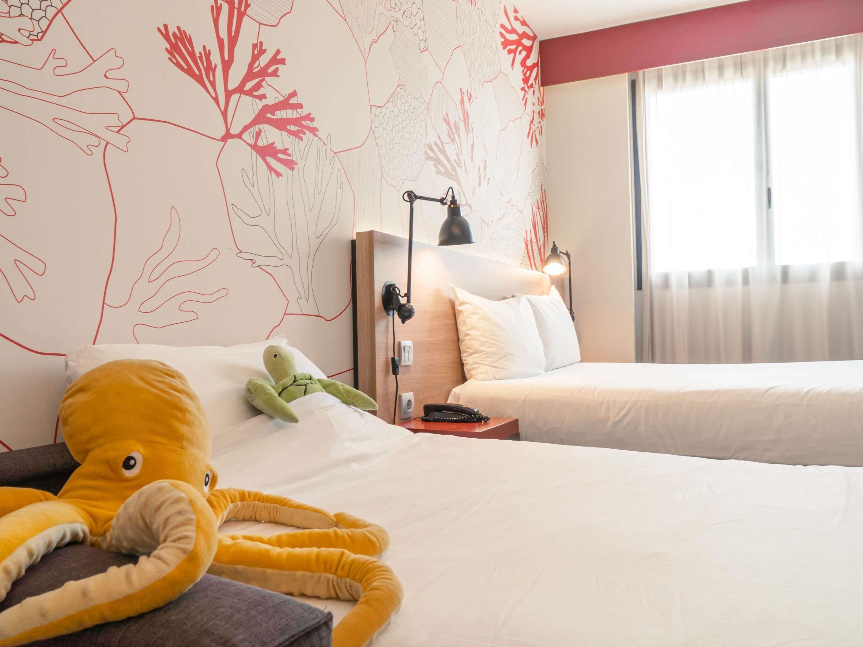Habitación de hotel con dos camas, un peluche de pulpo en la cama de la izquierda, una lámpara en la mesita de noche y una pared con estampado de arrecife de coral.