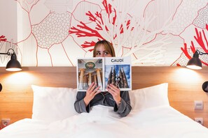 A person lying on a bed in a hotel room, holding a book titled "GAUDI". The room has a wooden headboard, two bedside lamps, and a wall with a red and white abstract design. - ibis Styles Barcelona City Bogatell (Barcelona)