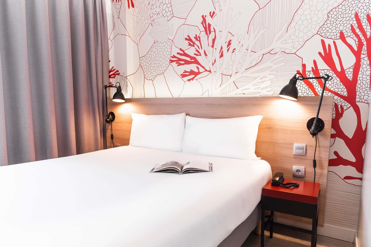A neatly made bed with a book, a bedside table with a lamp, and a wall with a tree pattern.