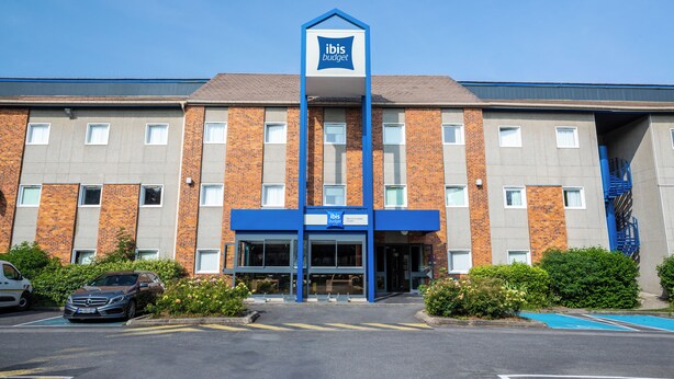 Un hôtel reconnaissable à son panneau « ibis budget » bien visible, à sa façade en briques et à son parking comprenant une place de stationnement bleue.