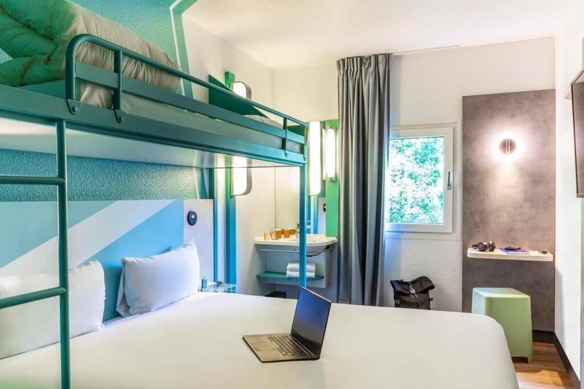 Habitación con literas, una computadora portátil sobre la cama inferior, un escritorio con una silla, una ventana con cortinas y un taburete pequeño.