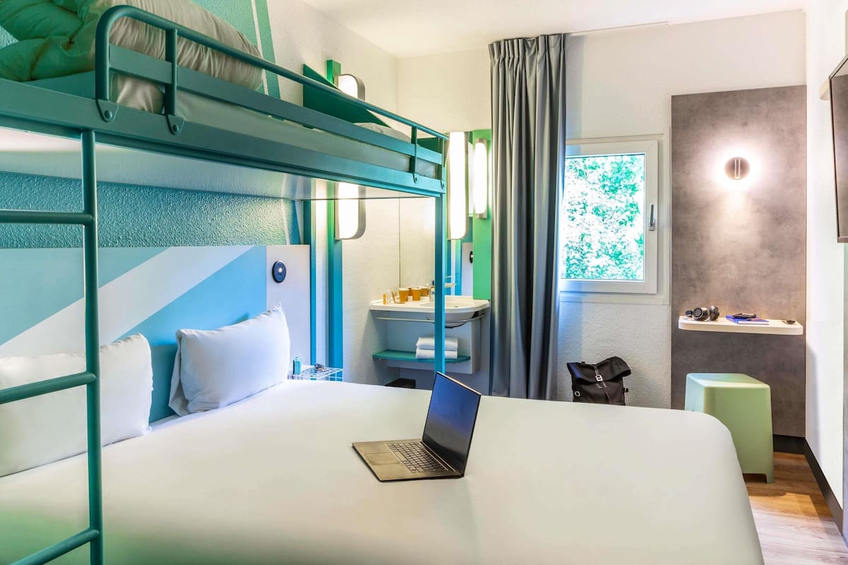 Un lit superposé avec un ordinateur portable posé sur le lit du bas, une table de chevet, une fenêtre avec des rideaux et un petit tabouret.