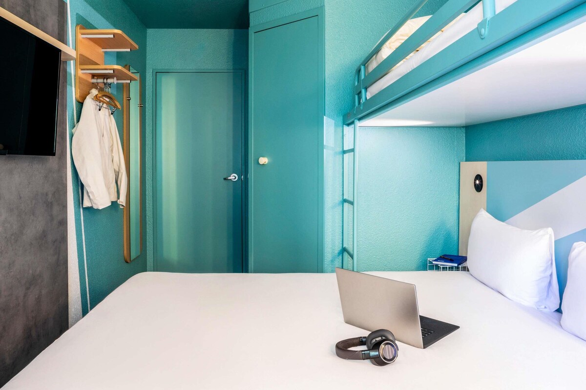 A room with bunk beds, a desk with a laptop and headphones, a TV, and a door with a coat rack.