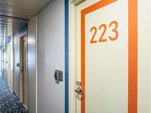 Pasillo de hotel con puertas numeradas 223, alfombra azul y un panel de control de seguridad.