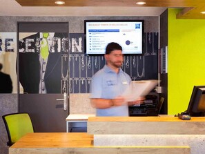 A reception desk with a digital display, a wooden counter, and a green chair. - ibis budget Marne la Vallee Chelles (Chelles)