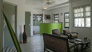 Una zona de recepción con un mostrador verde, sillas de mimbre, un jarrón grande y un reloj en la pared.