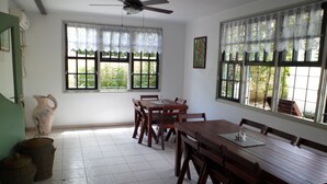 Un comedor con mesa y sillas de madera, ventanas amplias con cortinas de encaje, un ventilador de techo y piso de baldosas.