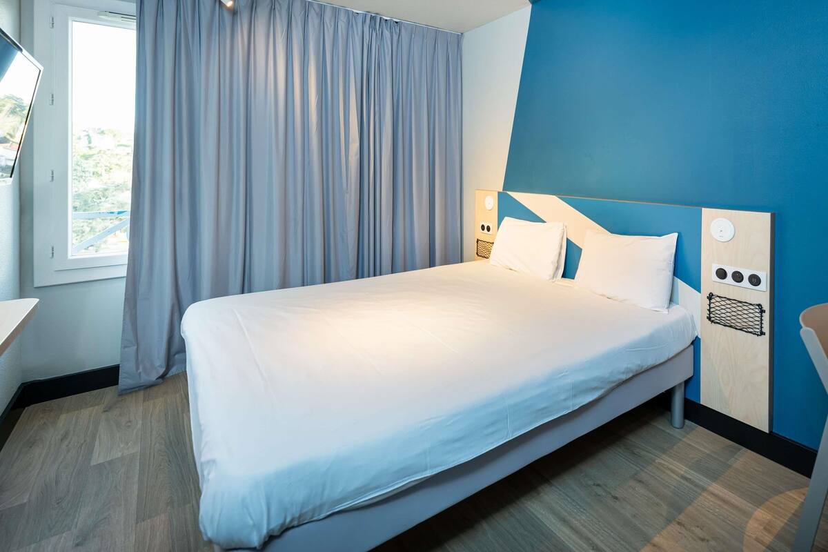 A neatly made bed in a room with a large window, a blue accent wall, and a wooden floor.