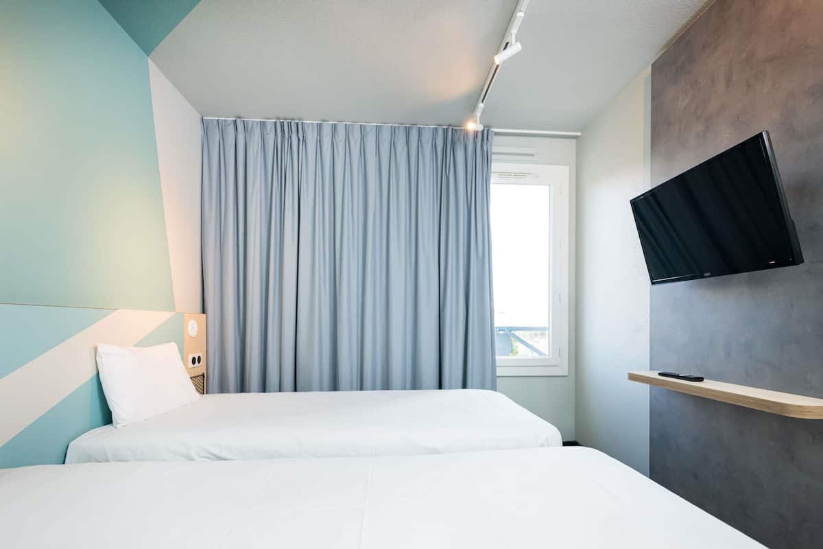 A hotel room with a bed, a TV mounted on the wall, a window with curtains, and a bedside table with a lamp.