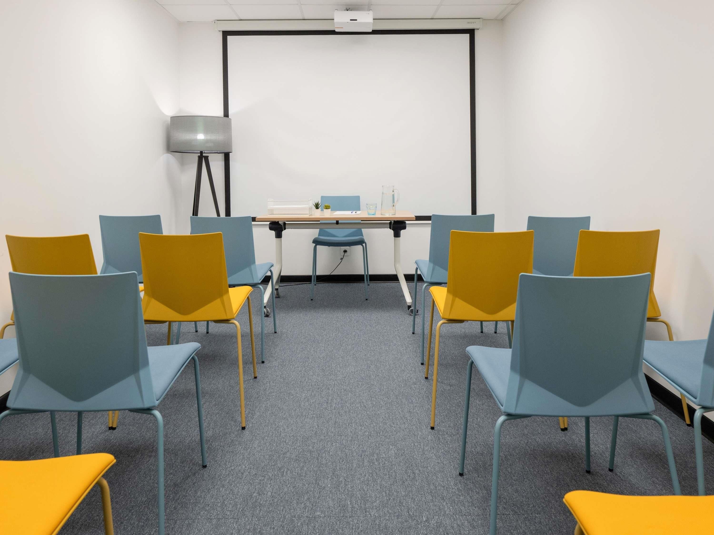 Une salle de conférence équipée d’un écran de projection, d’une table avec des chaises et d’une lampe.