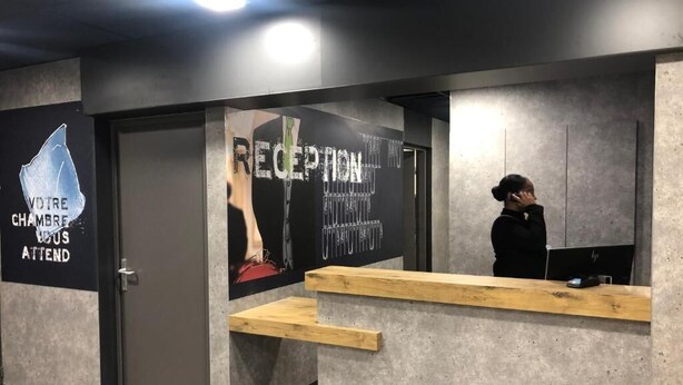 A reception area with a reception desk, a wall with a poster, and a door. - ibis budget Lyon Caluire Cite Internationale (Caluire-et-Cuire)