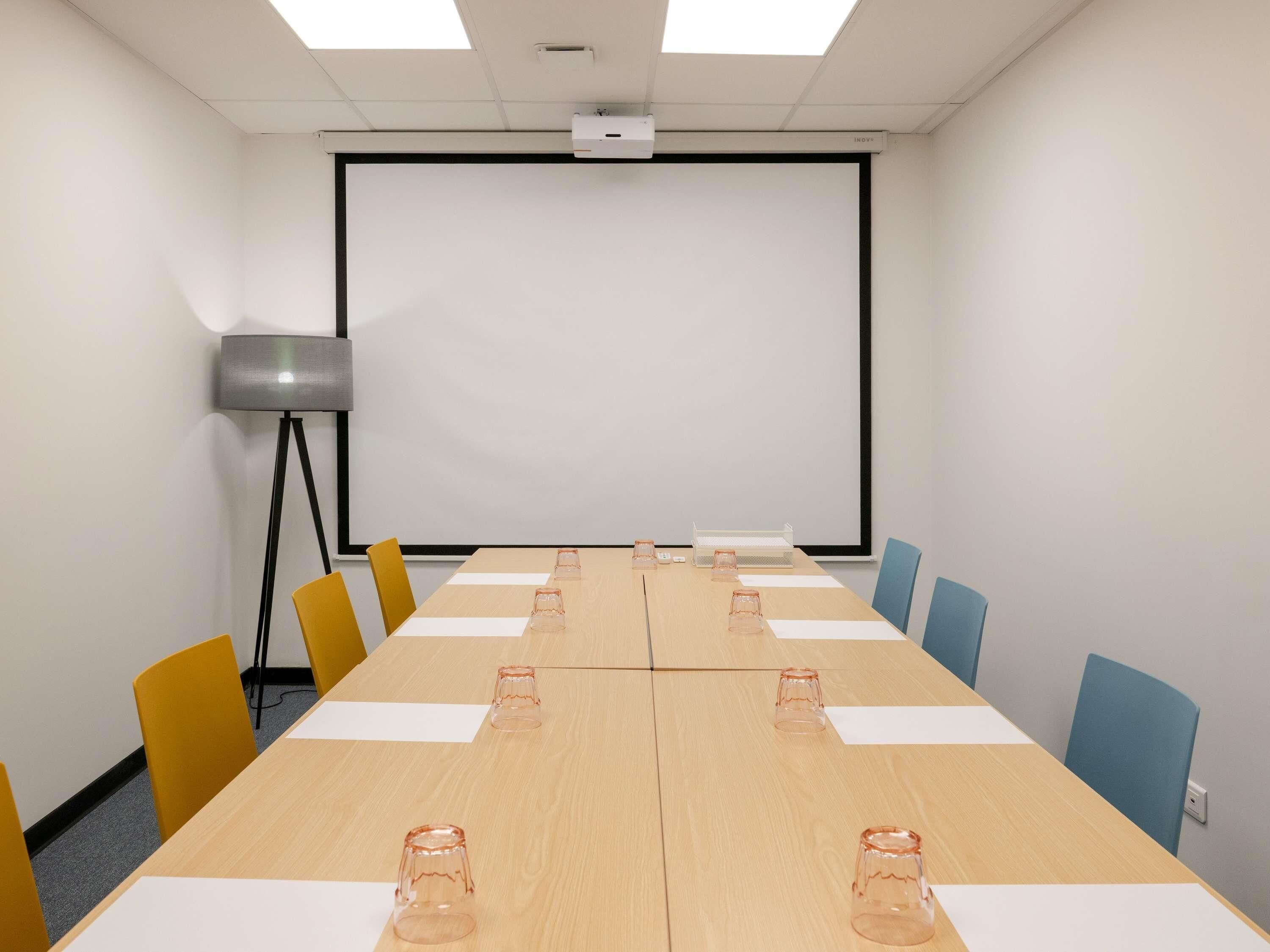 A conference room with a long table, chairs, a projector screen, and a floor lamp.