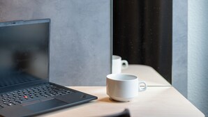 A laptop on a wooden table with a white mug beside it, near a window with a curtain. - ibis budget Lyon Caluire Cite Internationale (Caluire-et-Cuire)