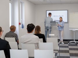 Un salón de conferencias con una pantalla de presentación que muestra 'HOGEO SALLES DE SECRETE', un orador en un atril y asistentes sentados mirando la pantalla.
