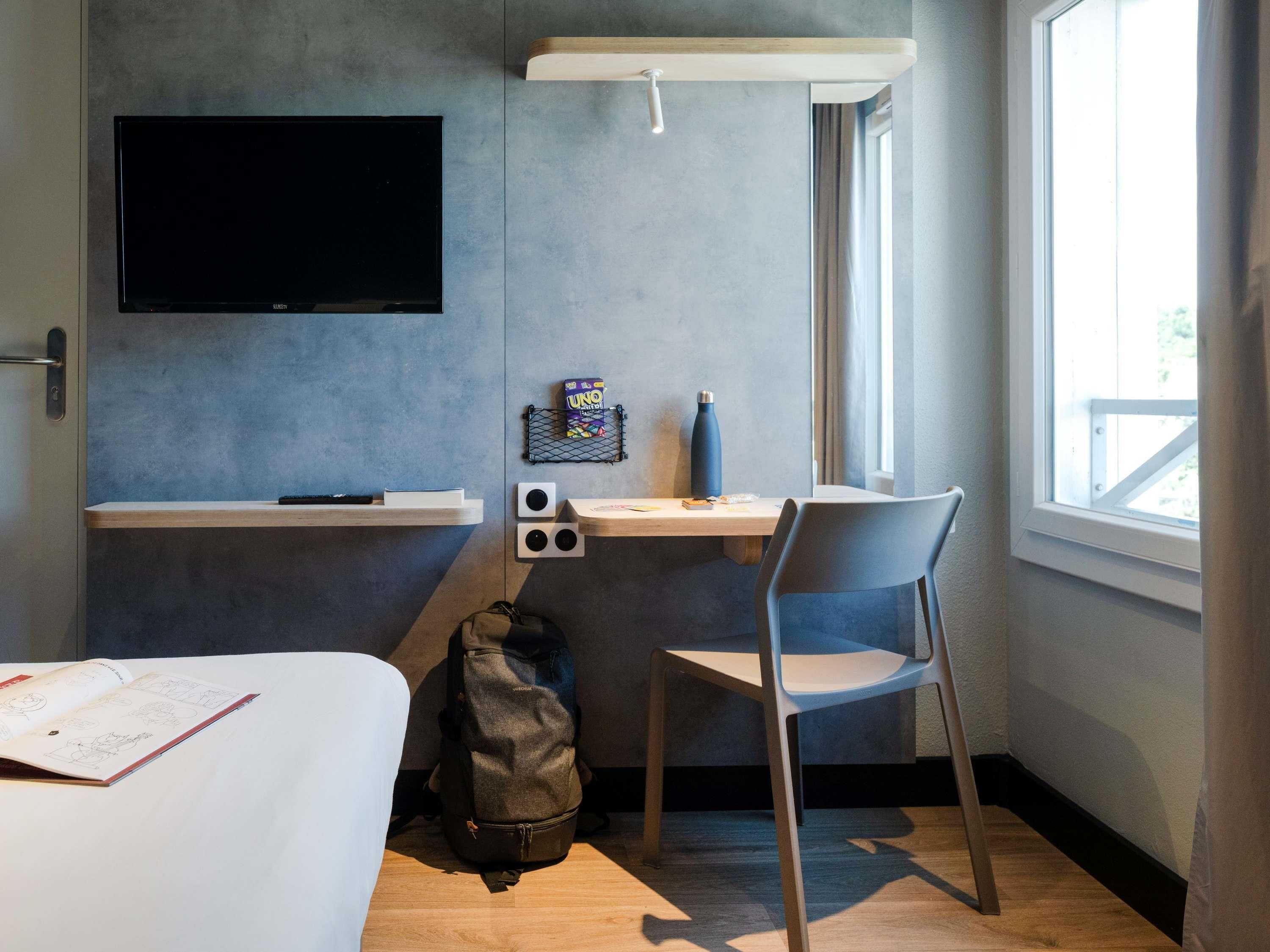 Habitación de hotel pequeña con cama, escritorio con silla, televisión en la pared y ventana que proporciona luz natural.