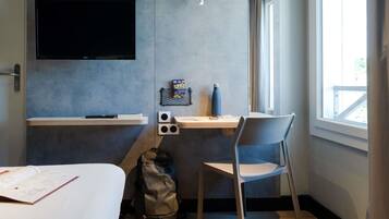 Habitación de hotel pequeña con cama, escritorio con silla, televisión en la pared y ventana que proporciona luz natural.