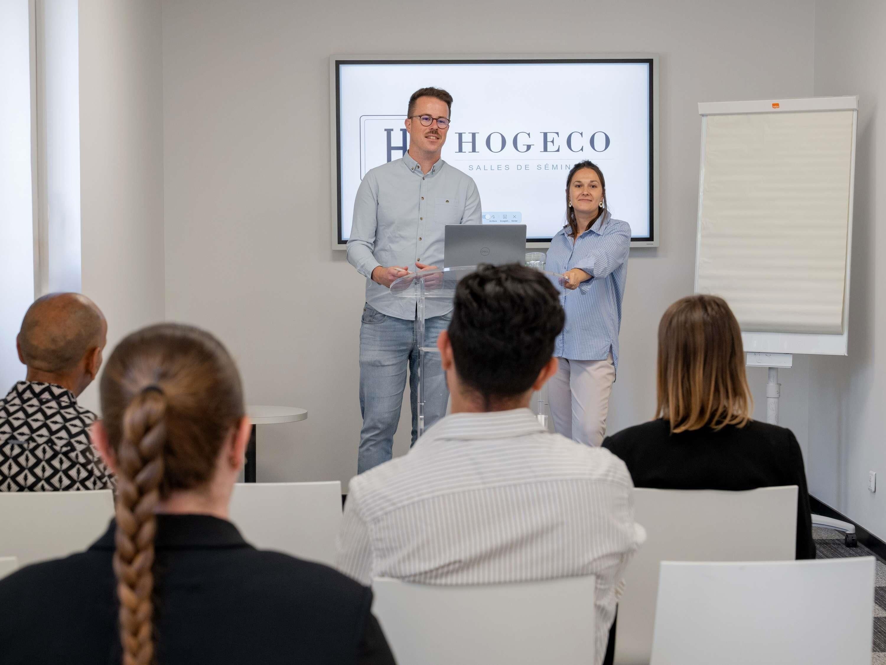 Una presentación en una sala de conferencias con una pantalla de proyector mostrando 'HOGEOCO' y 'SALLE DE SEMINAR'.