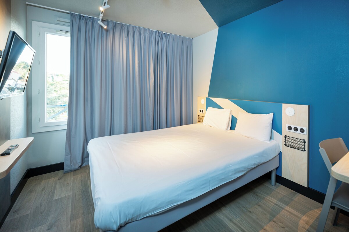 A modern hotel room with a large bed, a desk with a computer, and a window with curtains.