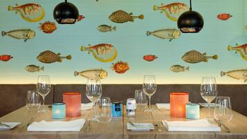 Ein Esstisch ist gedeckt mit Weingläsern, Kerzen und Besteck, an der Wand befinden sich Illustrationen von Fischen.