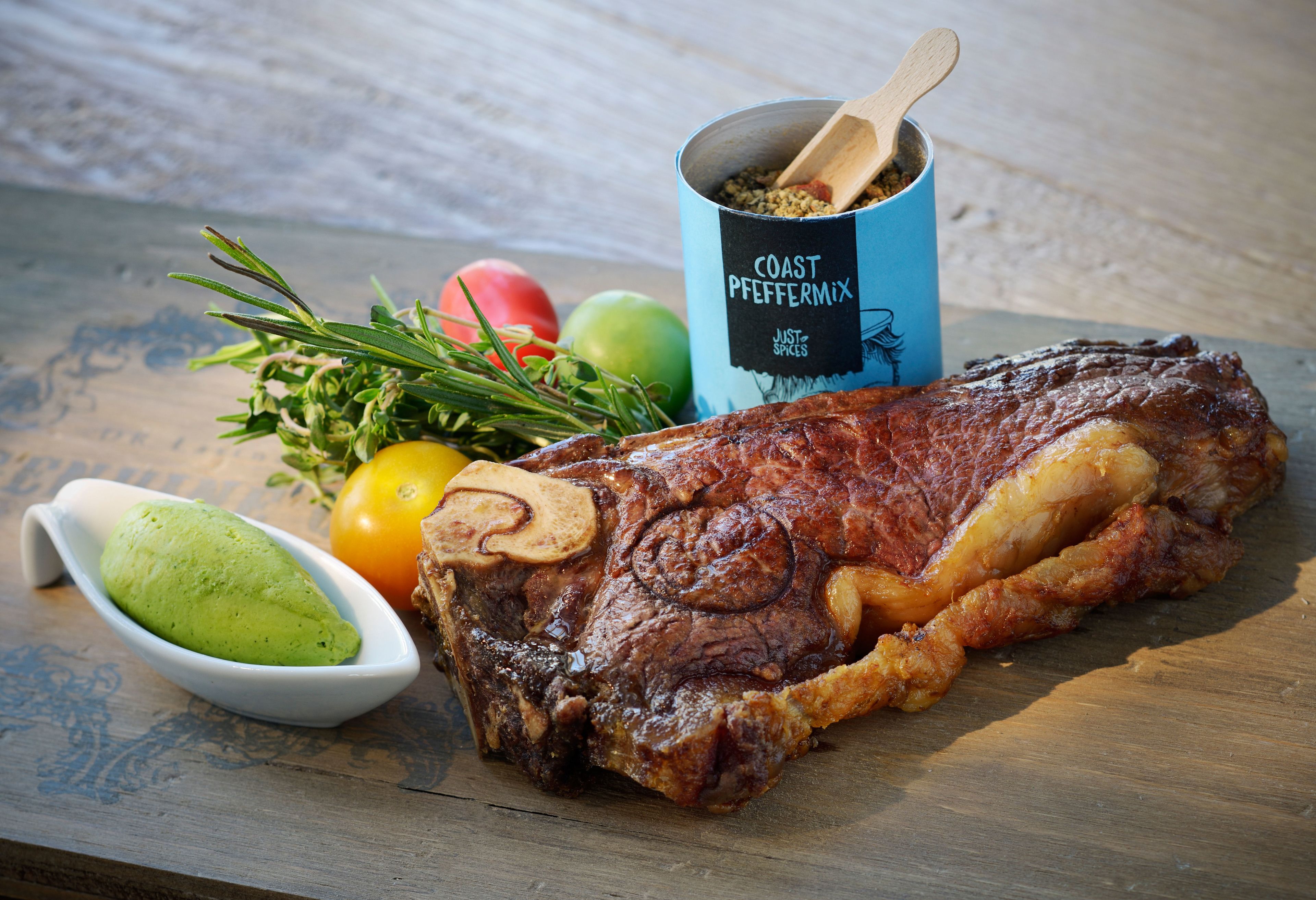 A steak with mushrooms, a bowl of green sauce, and a can of spices on a wooden surface.