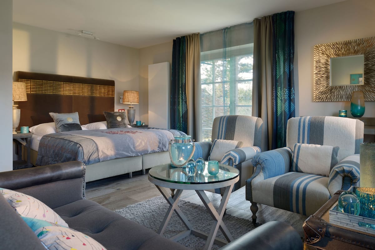 Ein modernes Hotelzimmer mit Bett, einer Couch, einem Sessel, einem Glastisch und einem Beistelltisch mit Dekoration.