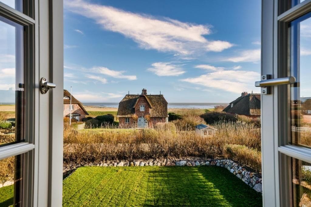 En ejendom ved kysten med græsplæne og stenkant, et stråtækt hus og en klar himmel.