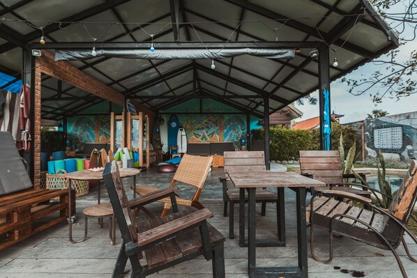 An outdoor seating area with wooden tables and chairs, a covered roof, and a mural on the wall. - Big Brother Surf Inn (Tibubeneng)