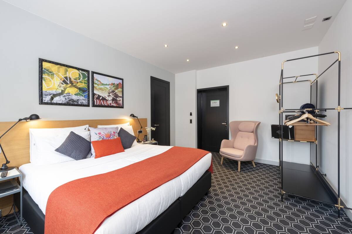 A modern hotel room with a neatly made bed, a chair, a wardrobe, and artwork on the walls.
