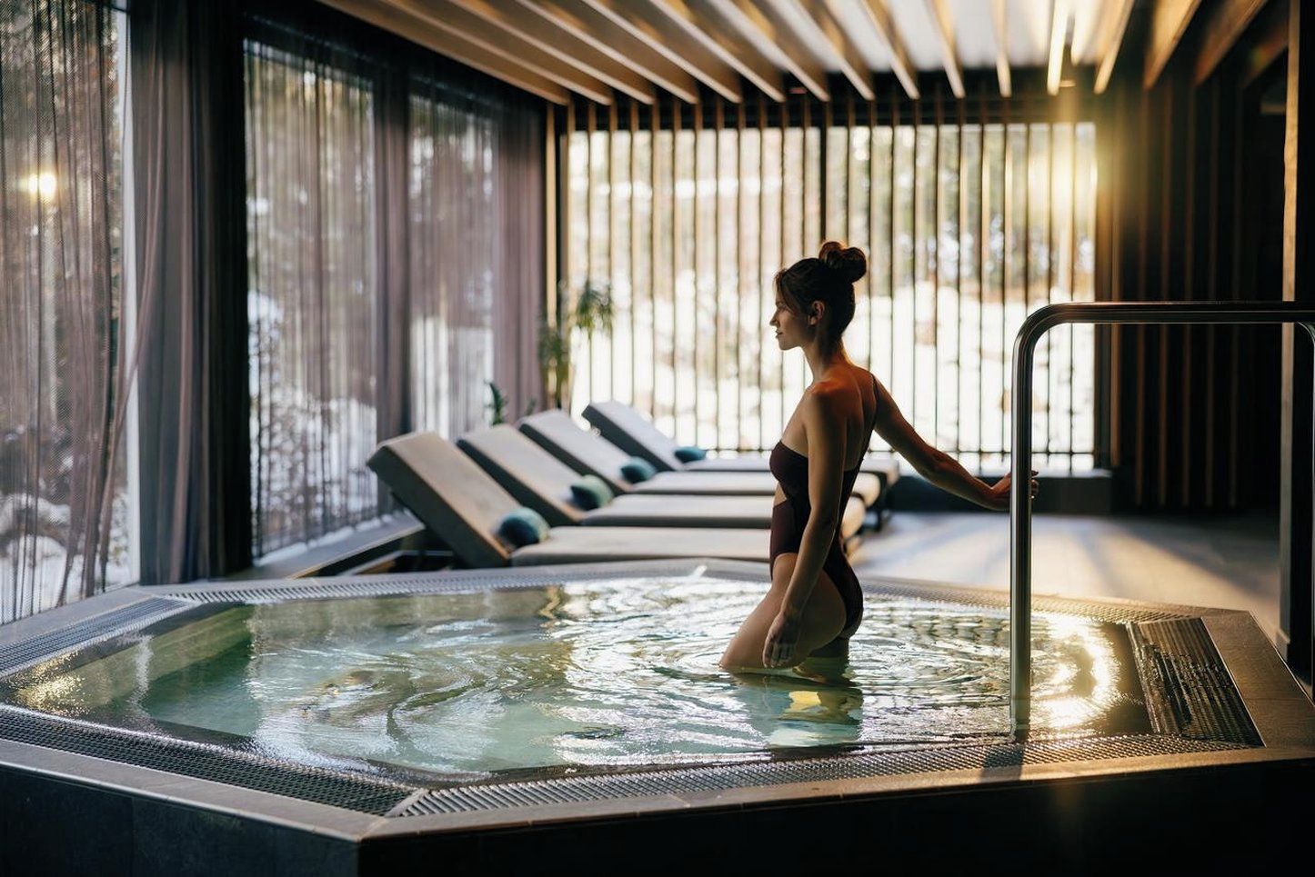 A person in a hot tub with a view of lounge chairs and a windowed wall.