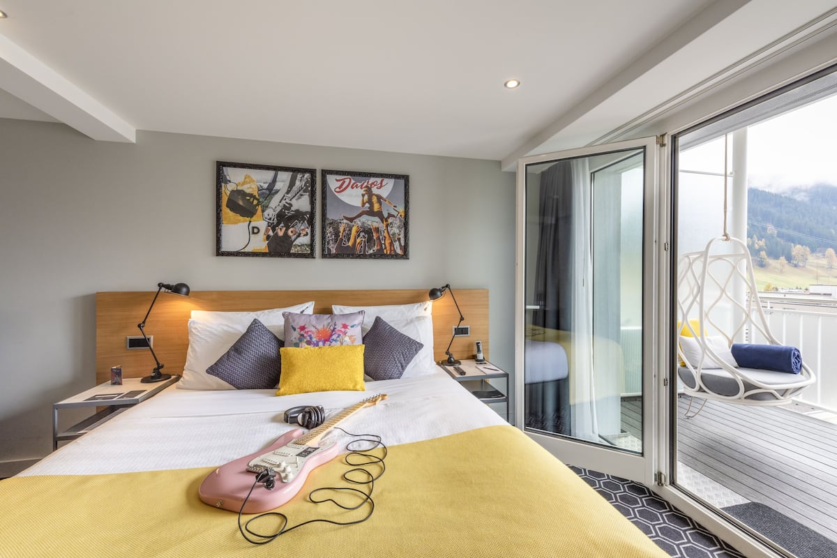 A modern hotel room with a large bed, a guitar, and a view of the mountains through the window.