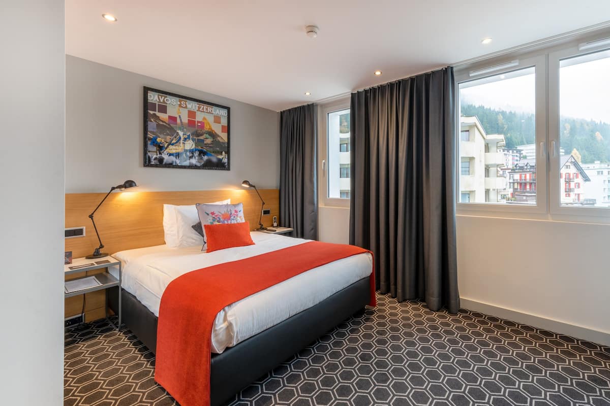 A hotel room with a large bed, a nightstand, a desk, a window with curtains, and a city view.