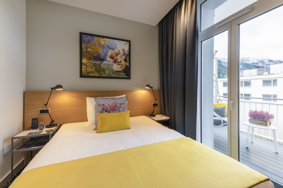 A hotel room with a bed, two bedside tables, a nightstand, a painting on the wall, and a view of the outside balcony.