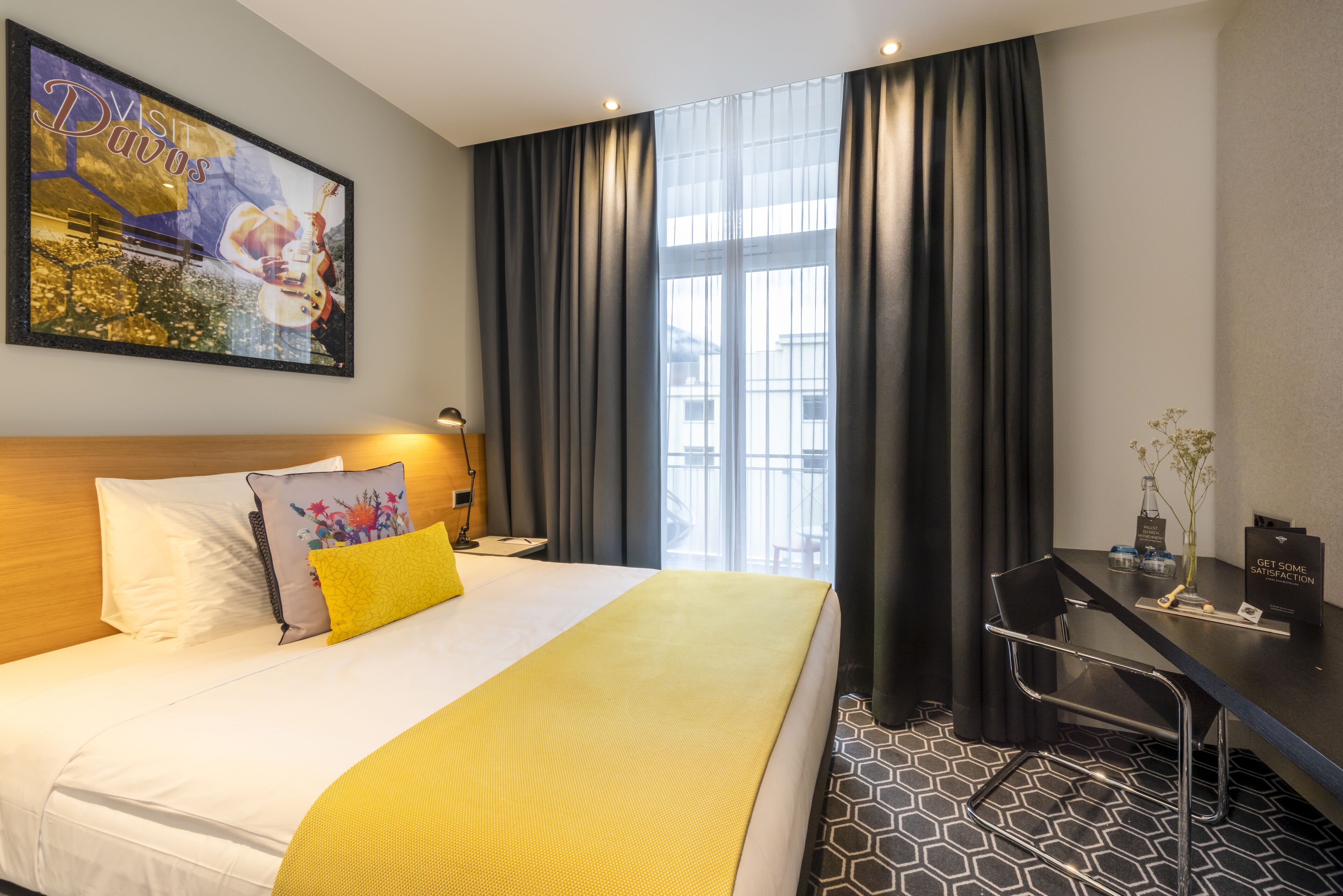 A hotel room with a bed, a desk, a chair, a nightstand, a lamp, a window with curtains, and a painting on the wall.