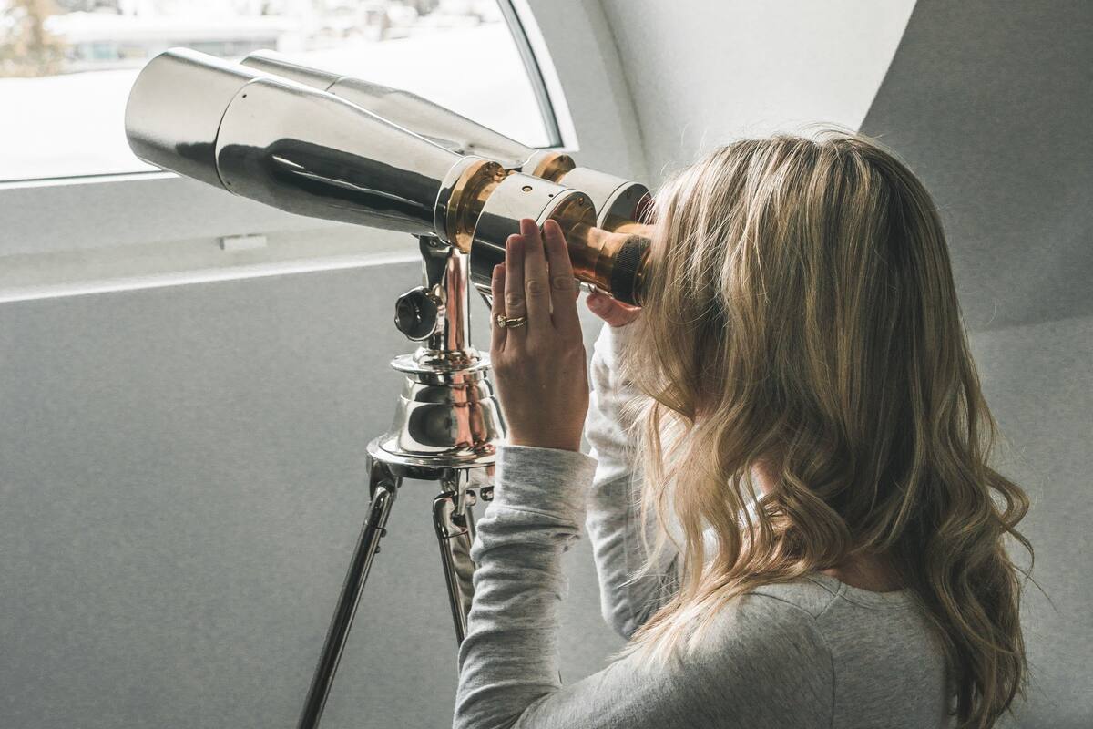 A person using a telescope inside a room with a curved ceiling and a window overlooking a snowy landscape.