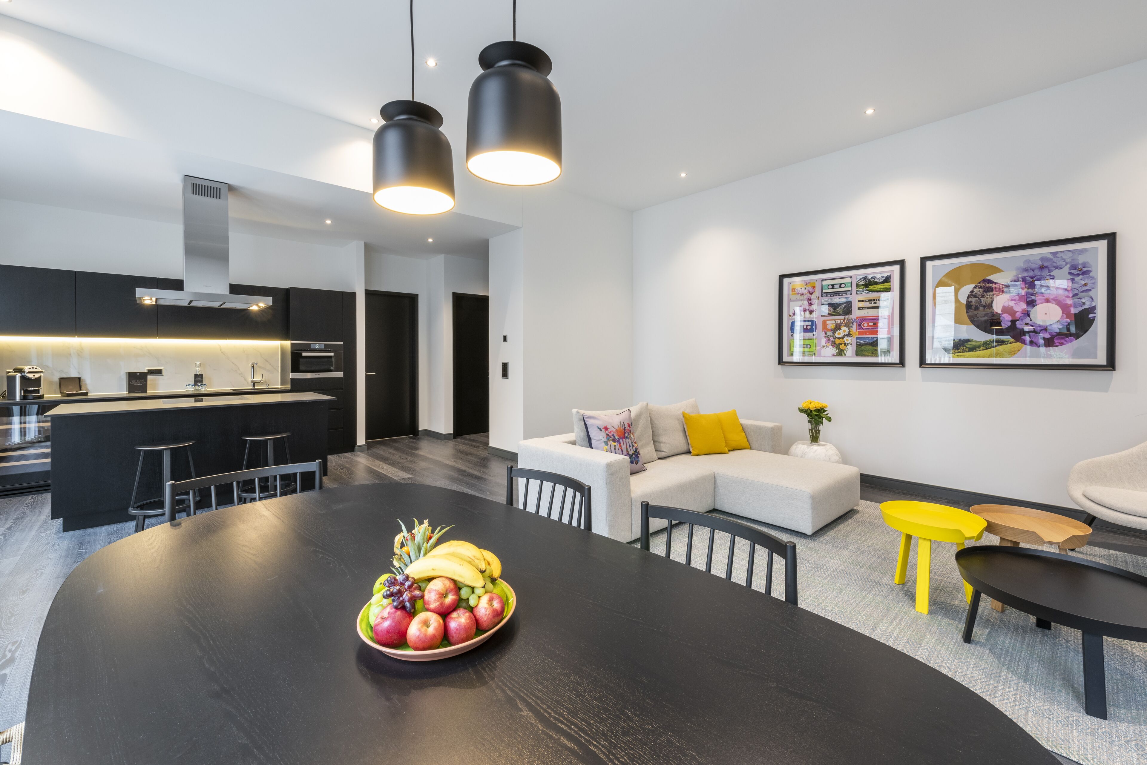 A modern kitchen with a central island, dining area with a black table and chairs, and a living area with a sofa and yellow side table.