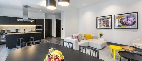 A modern kitchen with a central island, dining area with a black table and chairs, and a living area with a sofa and yellow side table.
