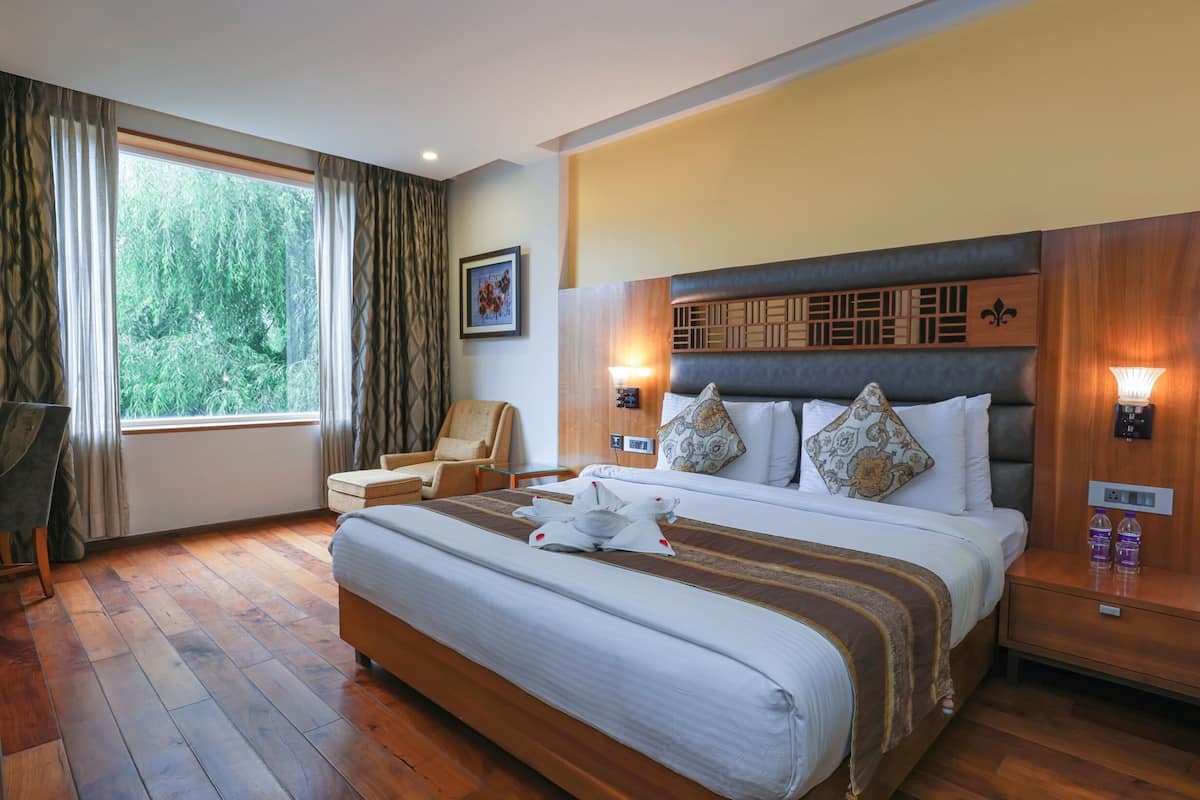 A hotel room with a large bed, wooden headboard, two bedside lamps, a chair, a small table, and a window with curtains.