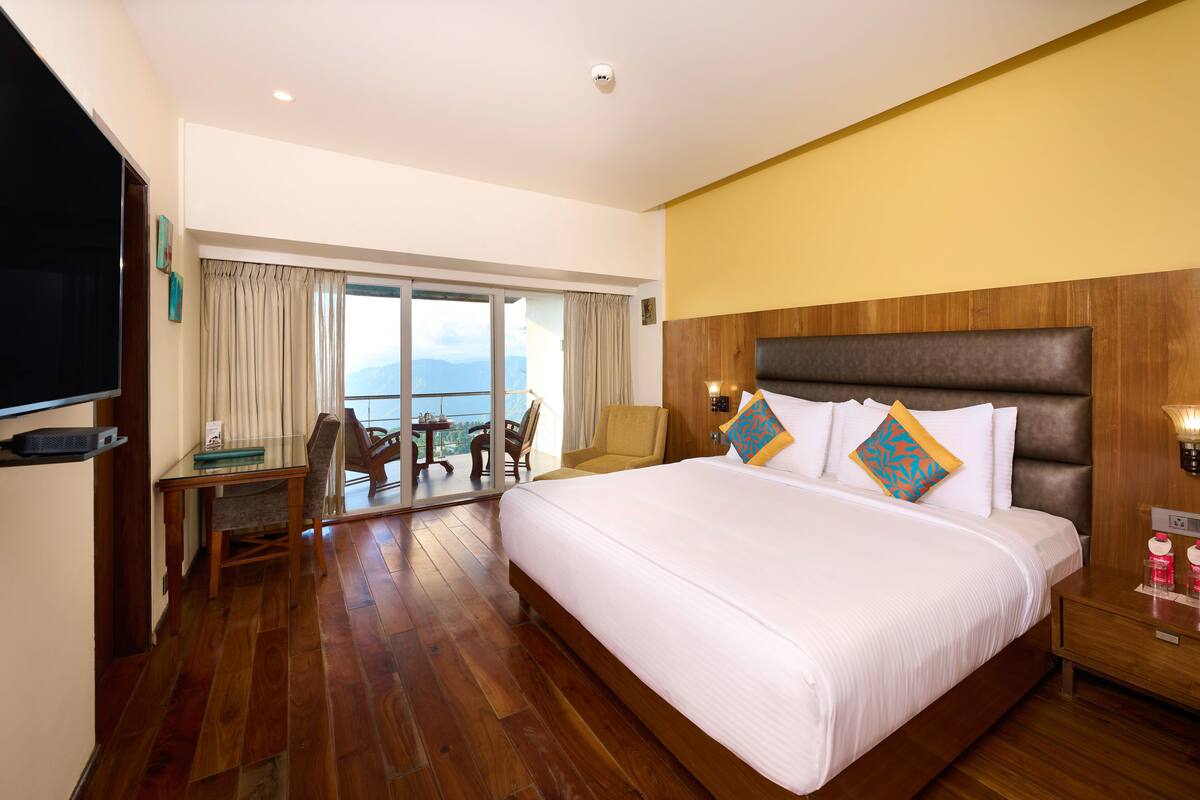 A hotel room with a large bed, a wooden headboard, a dining table with chairs, and a view of the sea through large windows.