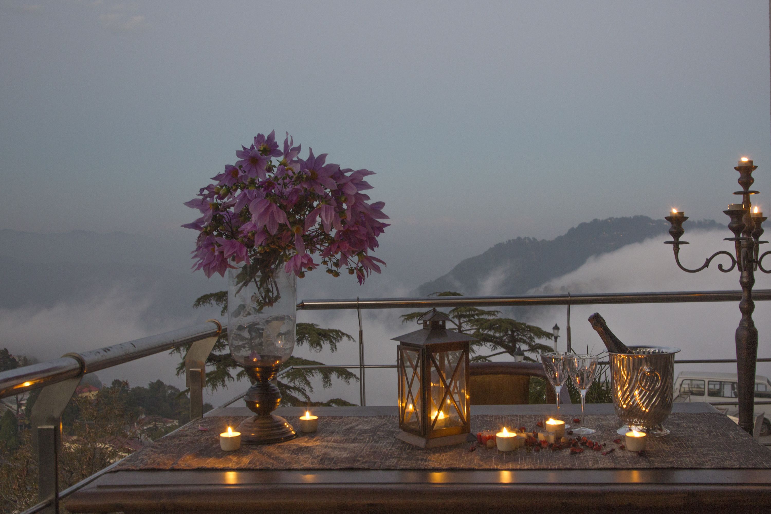 A table with a vase of flowers, candles, a lantern, and a bottle of champagne on a balcony overlooking a mountainous landscape.
