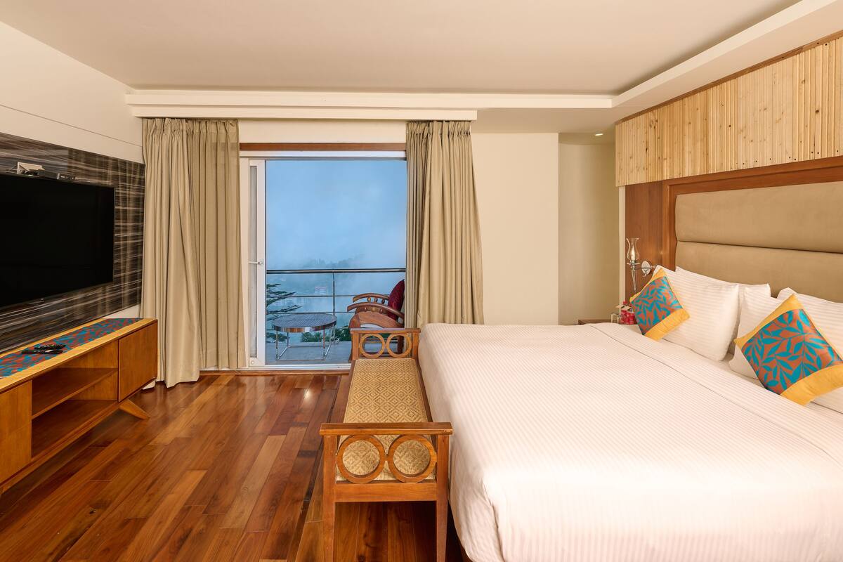 A hotel room with a large bed, a TV, a balcony with a view, and a wooden floor.