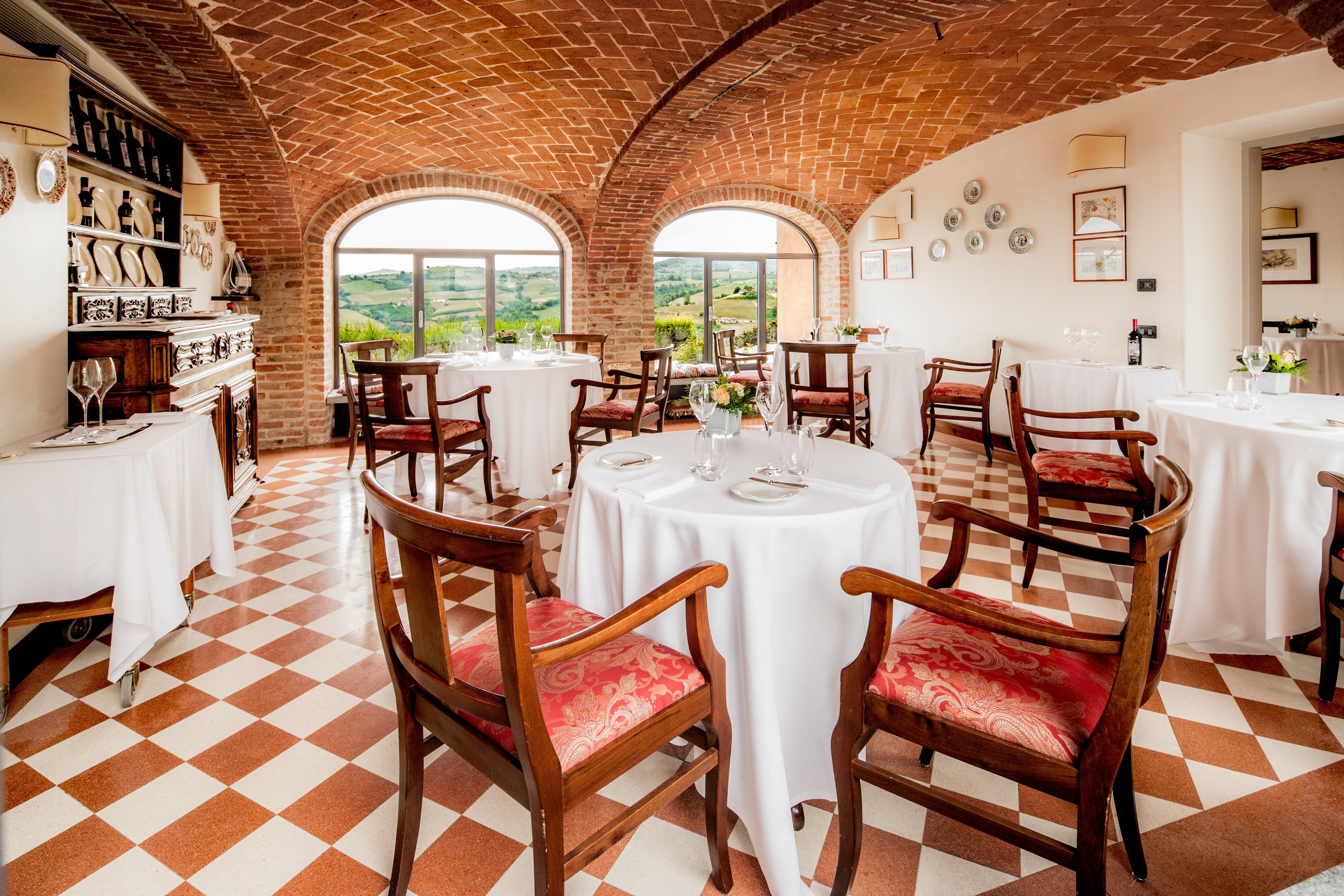 A restaurant with checkered flooring, wooden chairs with red cushions, white tables, and a brick ceiling.