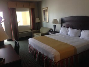 A hotel room with a large bed, a desk, a chair, a nightstand, a lamp, and a window with curtains.