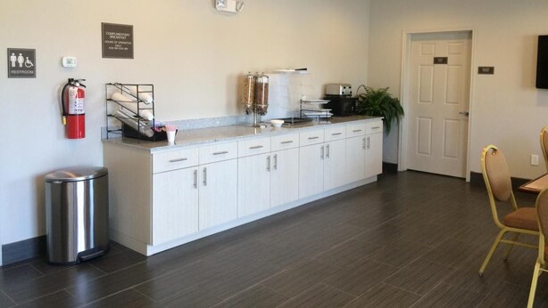 A clean, modern kitchen area with a stainless steel trash can, a coffee machine, and a microwave. There is a sign indicating breakfast hours and a fire extinguisher mounted on the wall. - Express Inn (Wharton)